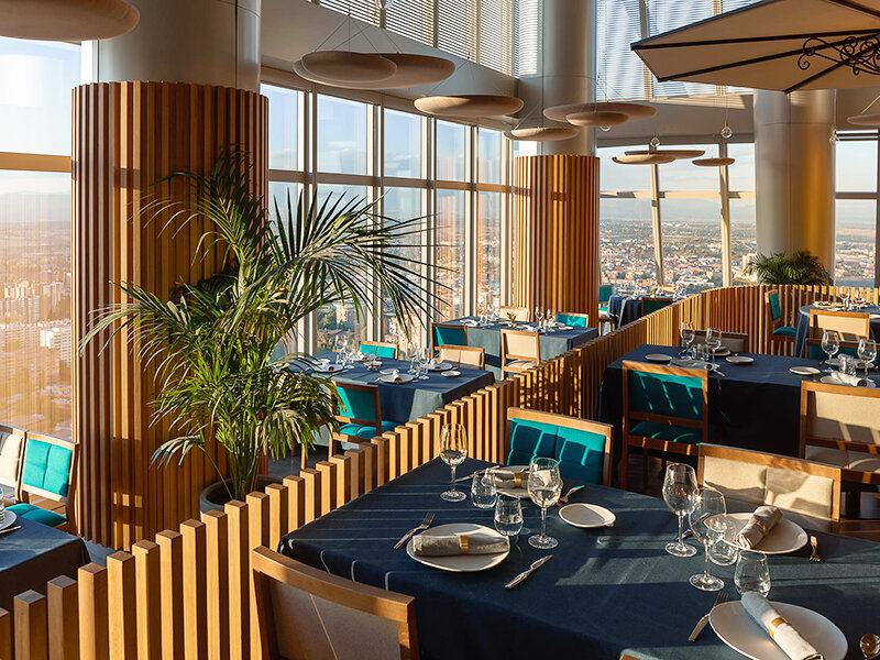 Un elegante restaurante con vistas a una ciudad durante el día, con detalles de madera y manteles azules.