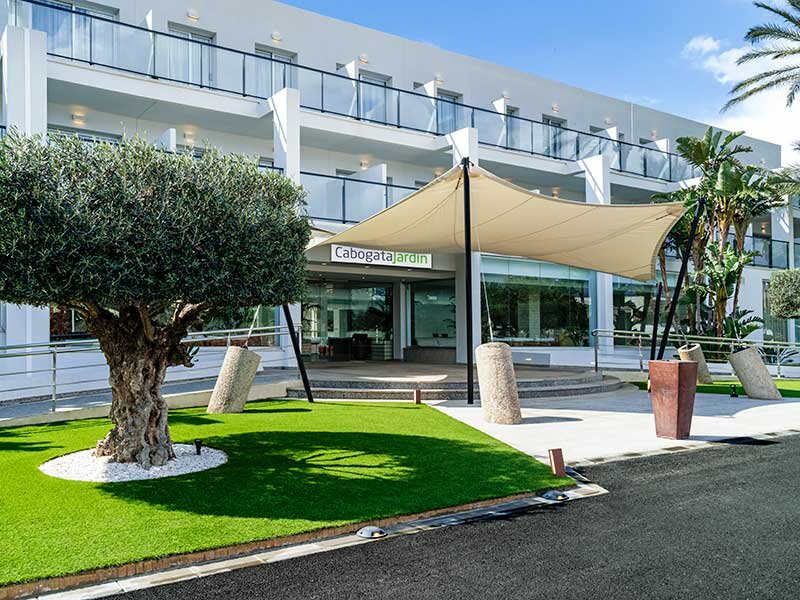 Entrada de hotel moderna con olivo grande, césped verde y toldo beige.