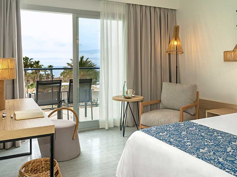Habitación de hotel con balcón con vistas al mar, palmeras, cama, silla, escritorio y lámparas.