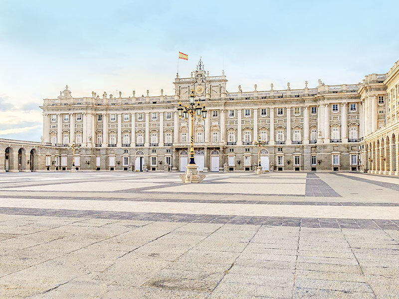 El Palacio Real de Madrid se alza majestuosamente bajo un cielo azul pálido con una bandera española ondeando.