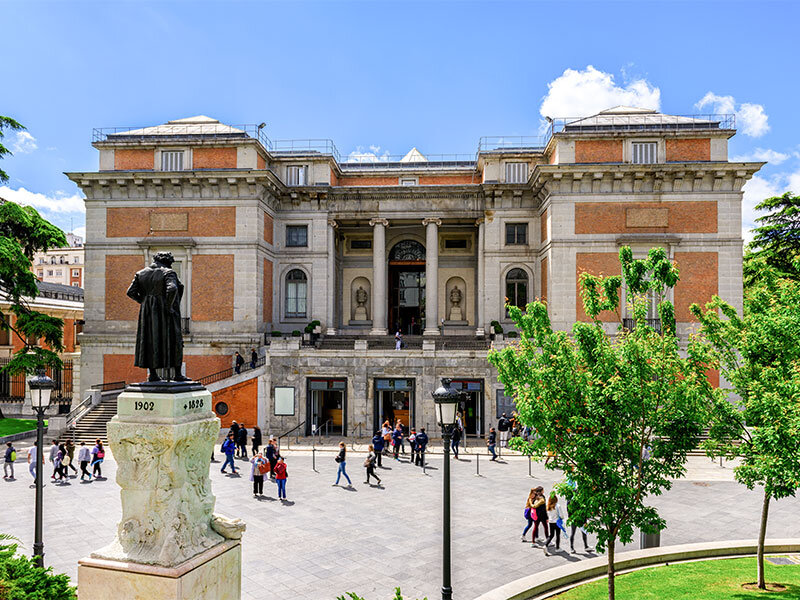 Una estatua se alza ante un edificio de ladrillo con columnas. Gente camina en una plaza.