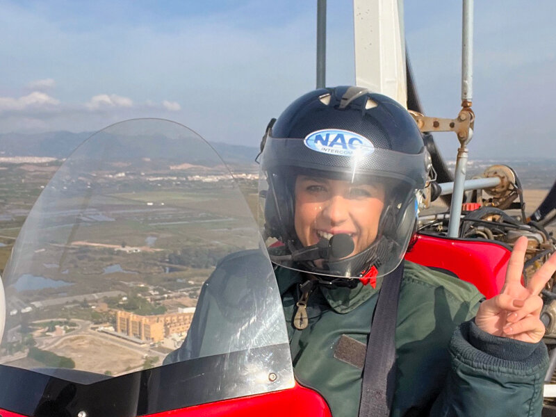 Mujer sonriente en casco hace signo de paz desde cabina de avioneta, sobre paisaje.
