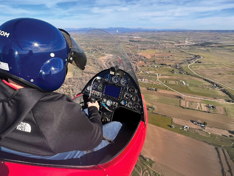 Piloto con casco azul volando sobre campos con cabina de avión en primer plano.