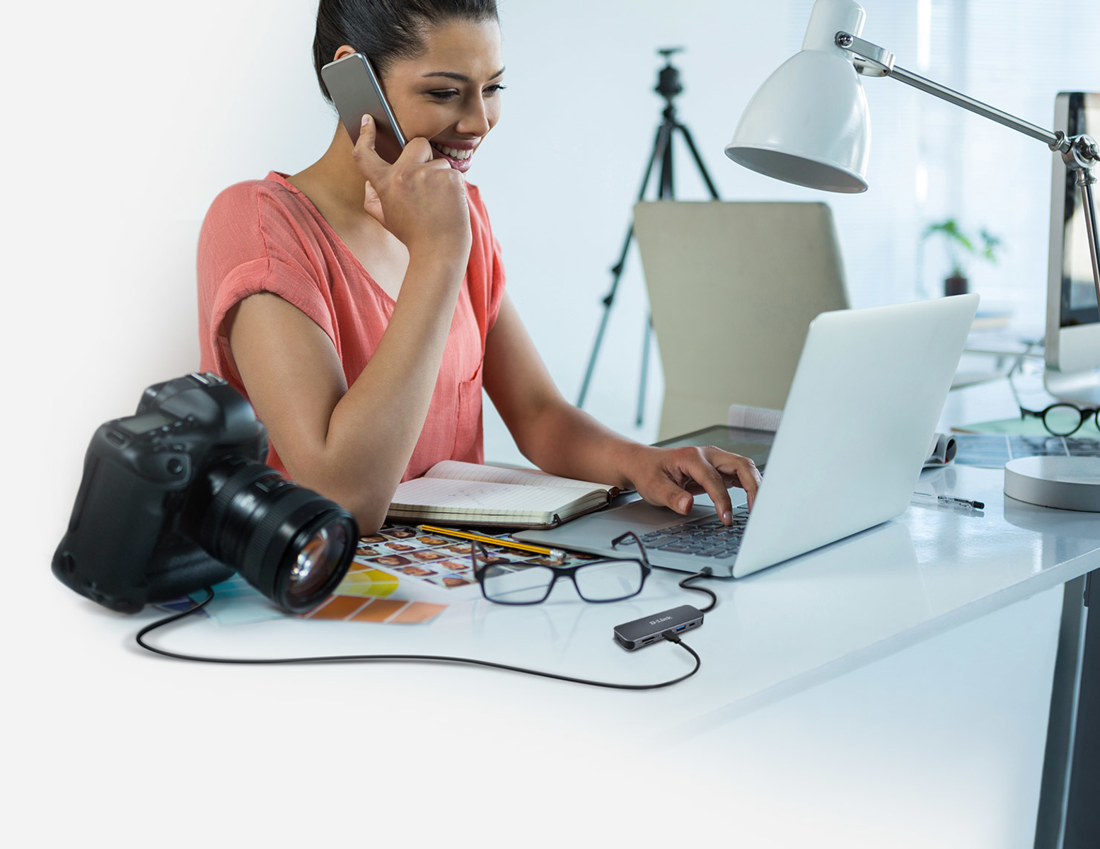 Una donna parla al telefono mentre digita su un laptop. Una fotocamera è vicina.