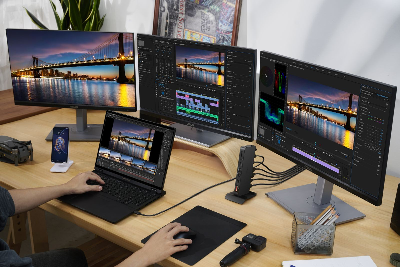 A person works on a laptop at a desk with three monitors displaying video editing software and a bridge scene.