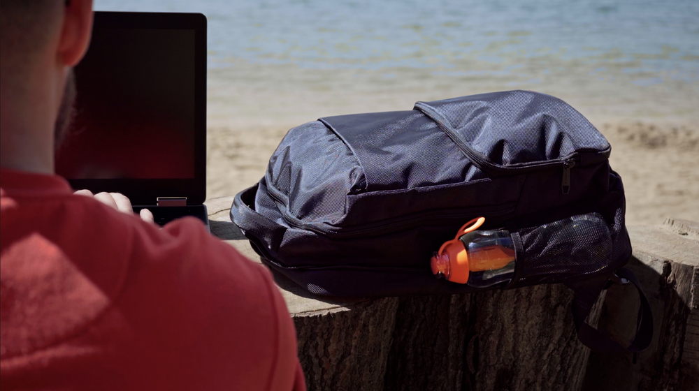 Mężczyzna w czerwonej koszuli pracuje na laptopie na plaży obok ciemnego plecaka i butelki z wodą.