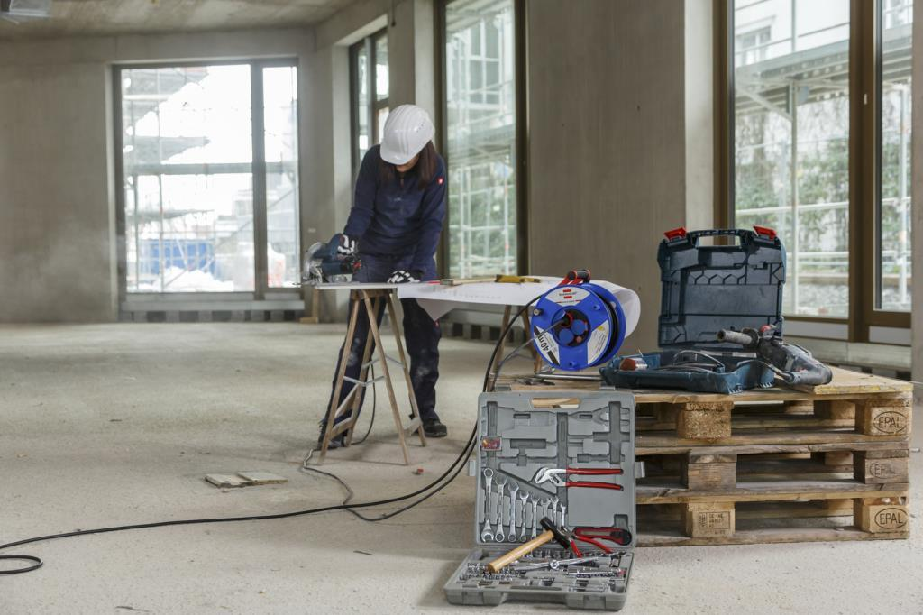 Bauarbeiterin sägt Holz auf Baustelle, Werkzeugkasten und Kabeltrommel.