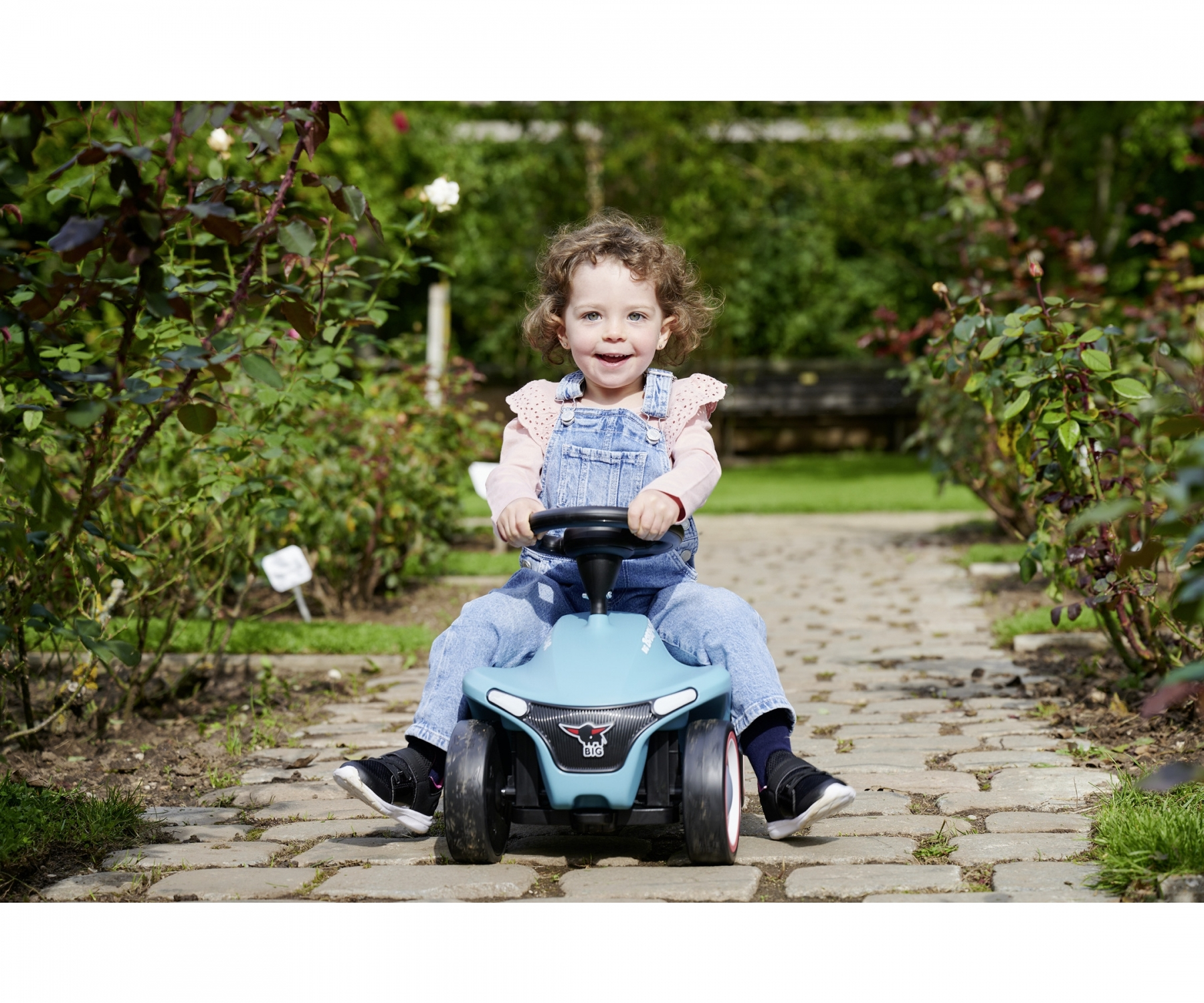 Ein Kleinkind sitzt auf einem blauen Spielzeugauto im Freien, umgeben von Rosenbüschen.