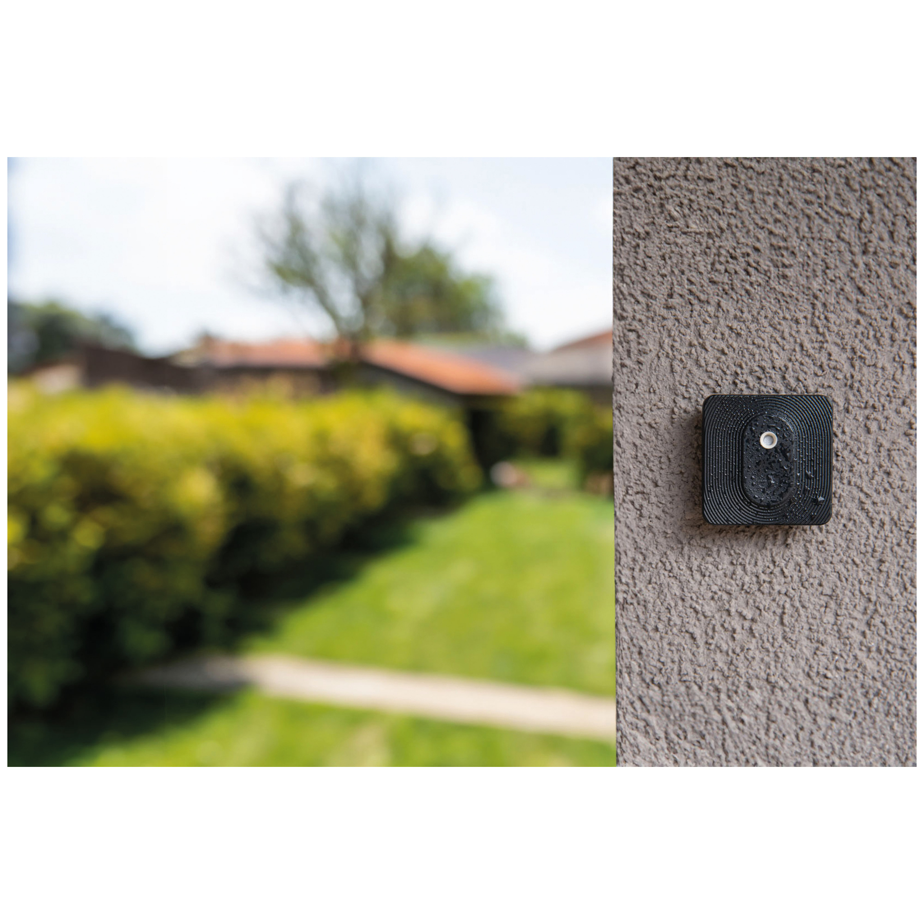 Schwarzer Sensor mit ovaler Linse an einer grauen, strukturierten Wand; Garten im Hintergrund.