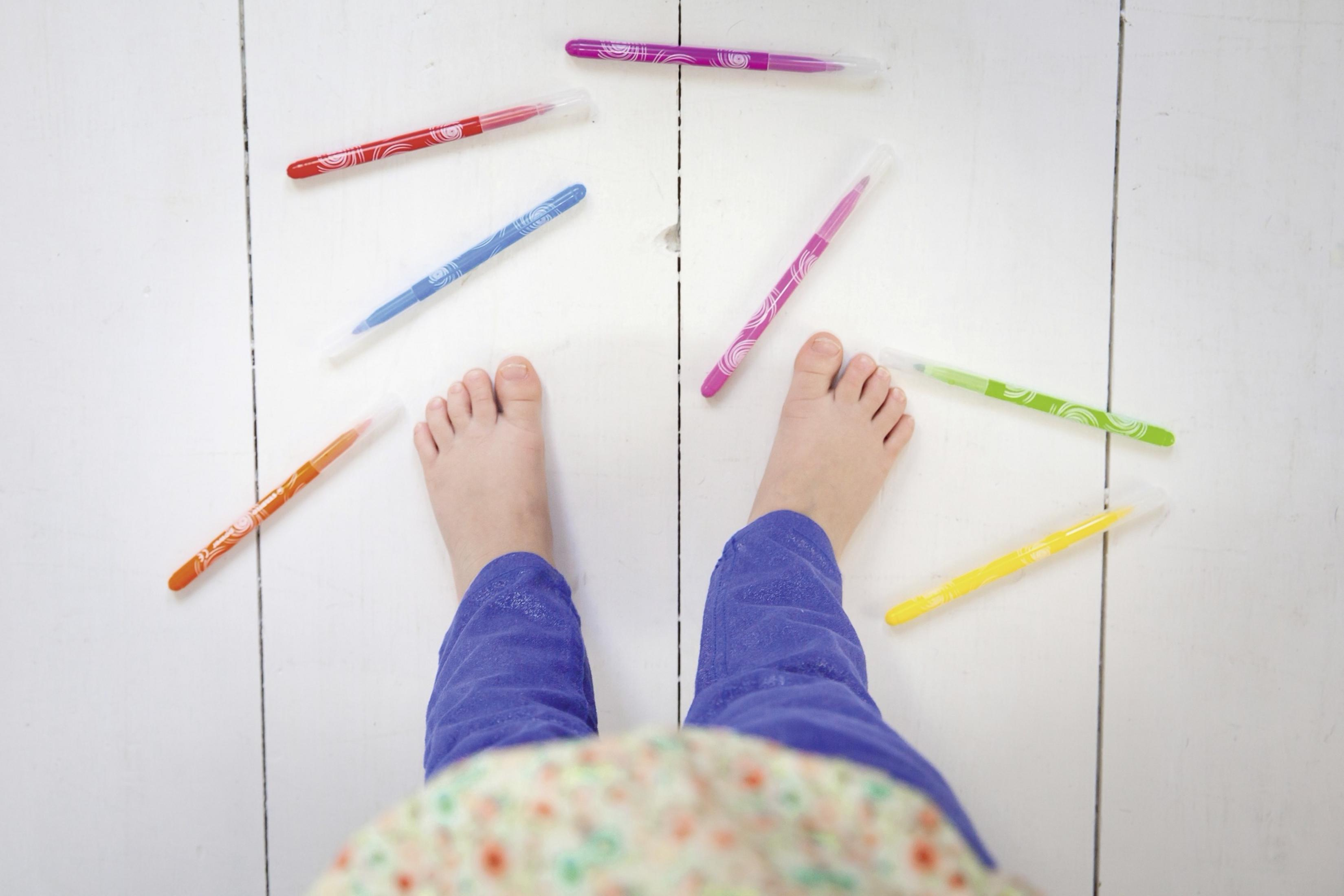 Kinderfüße umgeben von bunten Stiften auf weißem Holzboden.