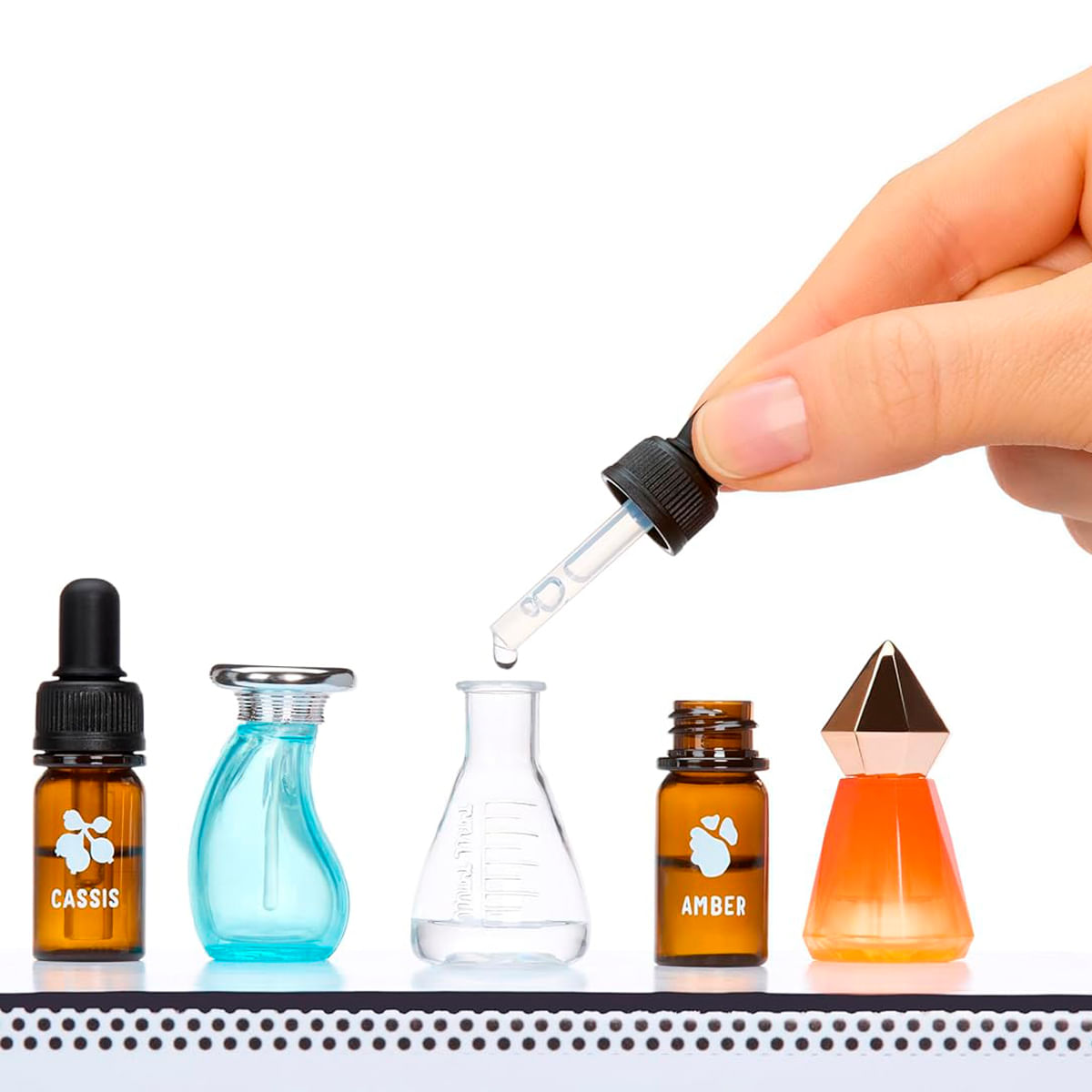 Una mano sostiene un cuentagotas sobre botellas pequeñas: ámbar 'Cassis', azul, matraz transparente, ámbar 'Ámbar', naranja.