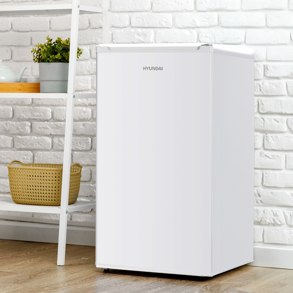 Un refrigerador blanco HYUNDAI junto a un estante blanco con cestas y plantas.