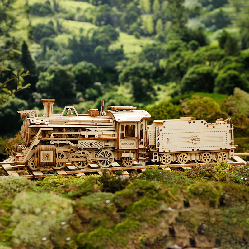 Ahşap bir model tren, yemyeşil bitki örtüsünün ortasında raylar üzerinde duruyor. Tren tekerlekler, borular ve tender vagon ile detaylandırılmış.