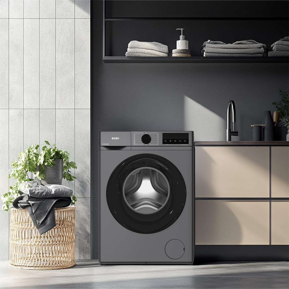 Lavadora gris con carga, al lado una cesta de ropa y una planta.