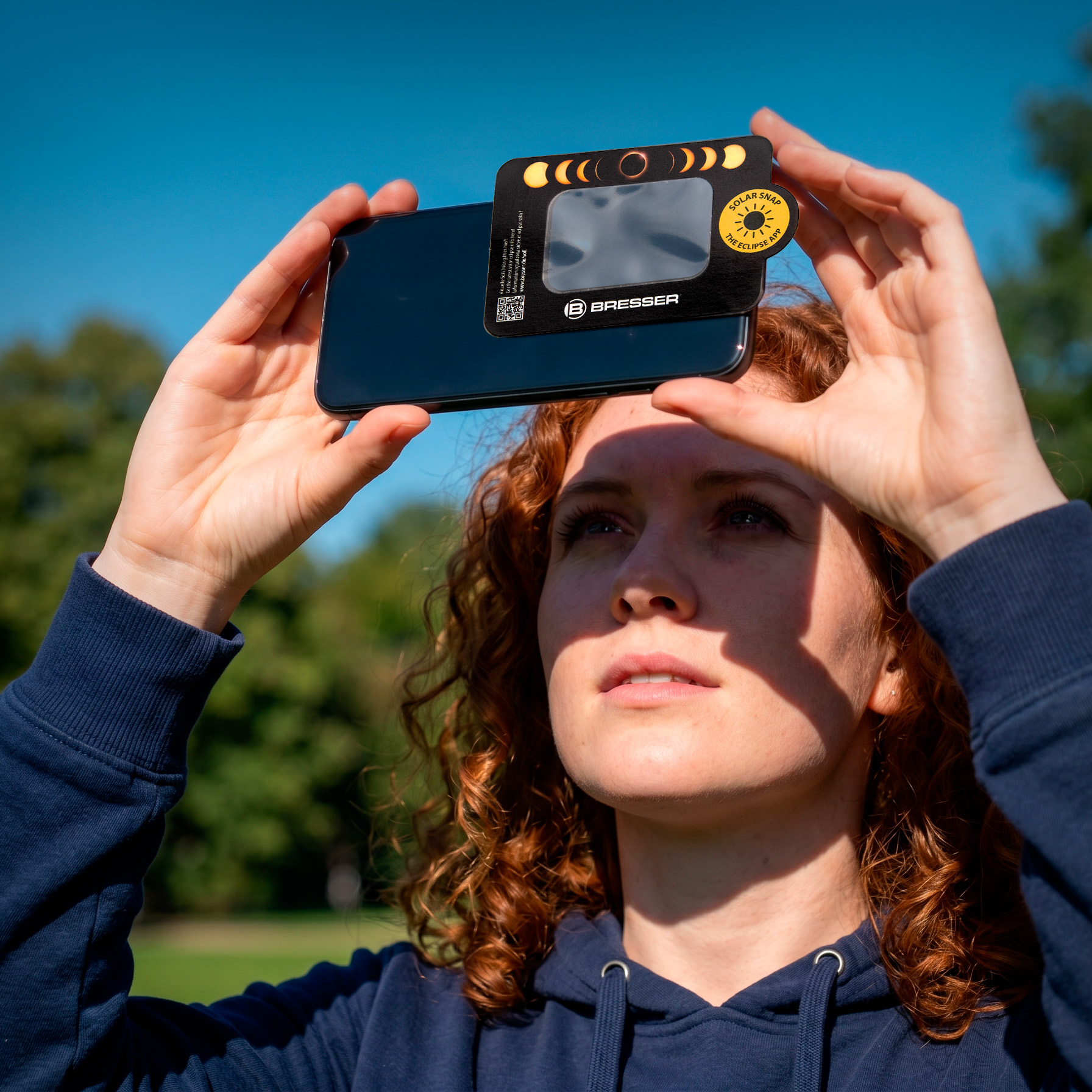 Una mujer pelirroja sostiene un teléfono inteligente con filtro solar frente al sol.