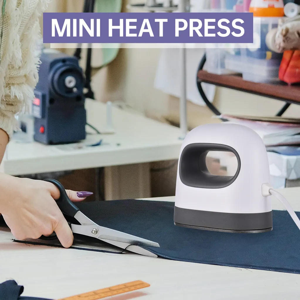 Weiße Mini-Heißpresse mit dunklem Griff auf dunkelblauem Stoff neben einer Schere.