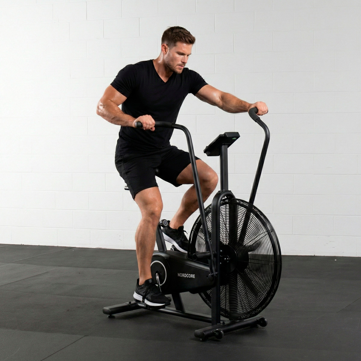 Ein Mann in Schwarz trainiert auf einem schwarzen Airbike vor einer weißen Ziegelwand.
