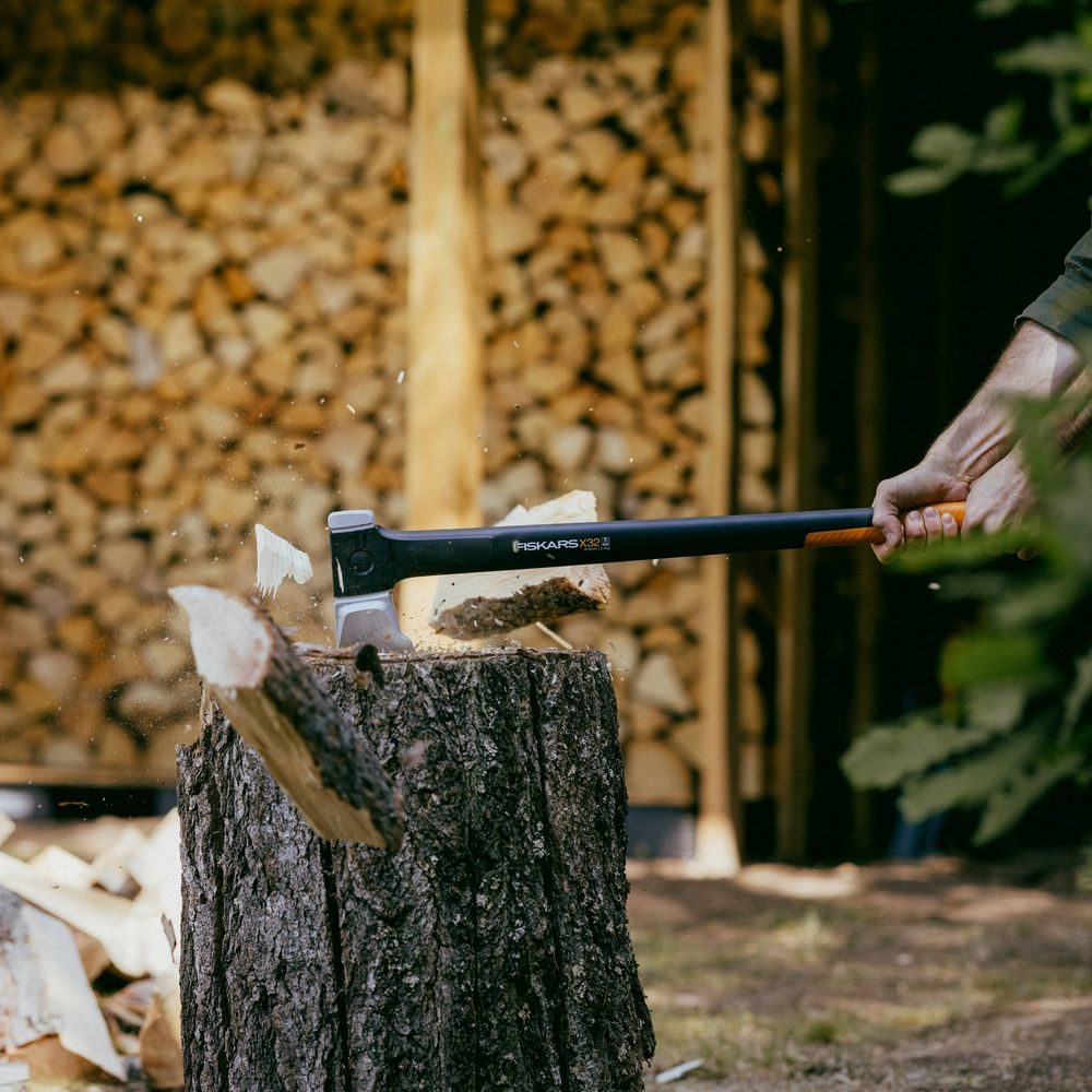 Mann spaltet Holz mit Axt auf Baumstumpf, Holzstapel im Hintergrund.