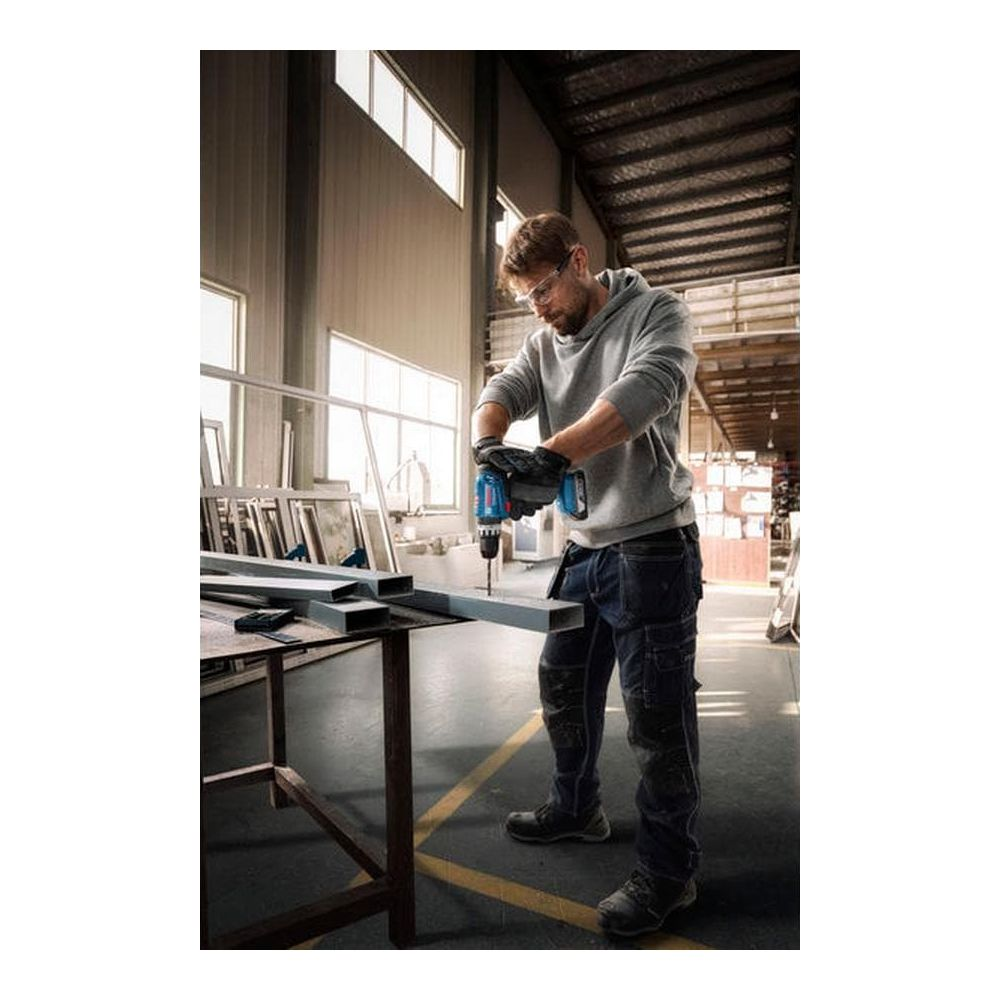 Mann bohrt in Metallprofil auf Werkbank, trägt Schutzbrille und Handschuhe.