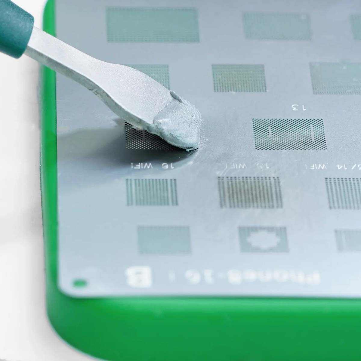 Eine Metallschablone mit Lötpaste, die mit einem Spatel auf eine grüne Oberfläche aufgetragen wird.