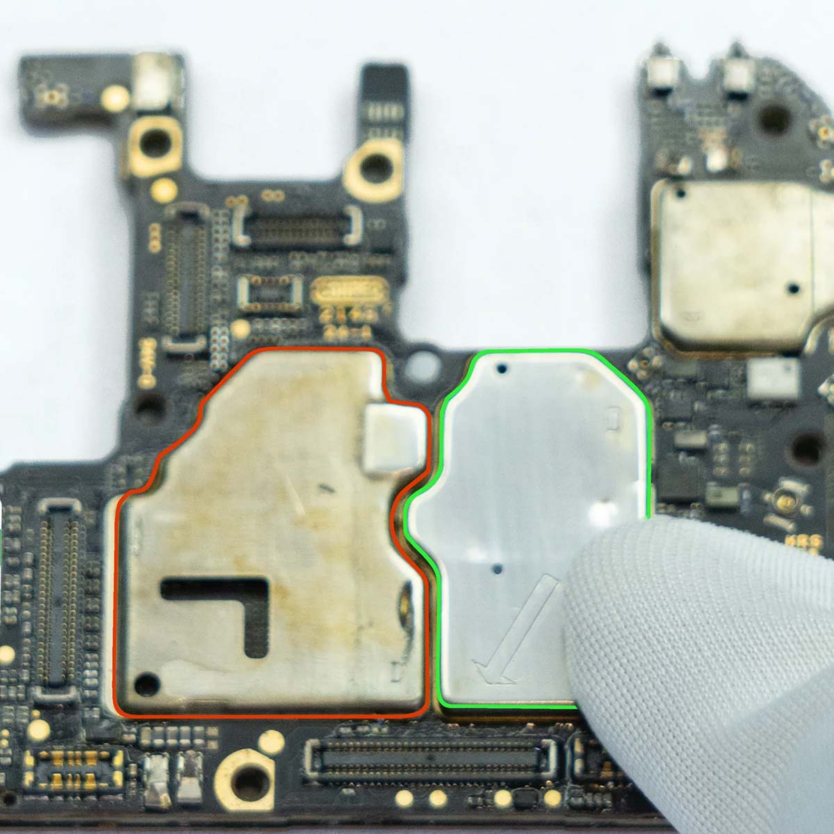 Primo piano di una scheda di circuito con due scudi metallici evidenziati. Una mano guantata tocca lo scudo destro.