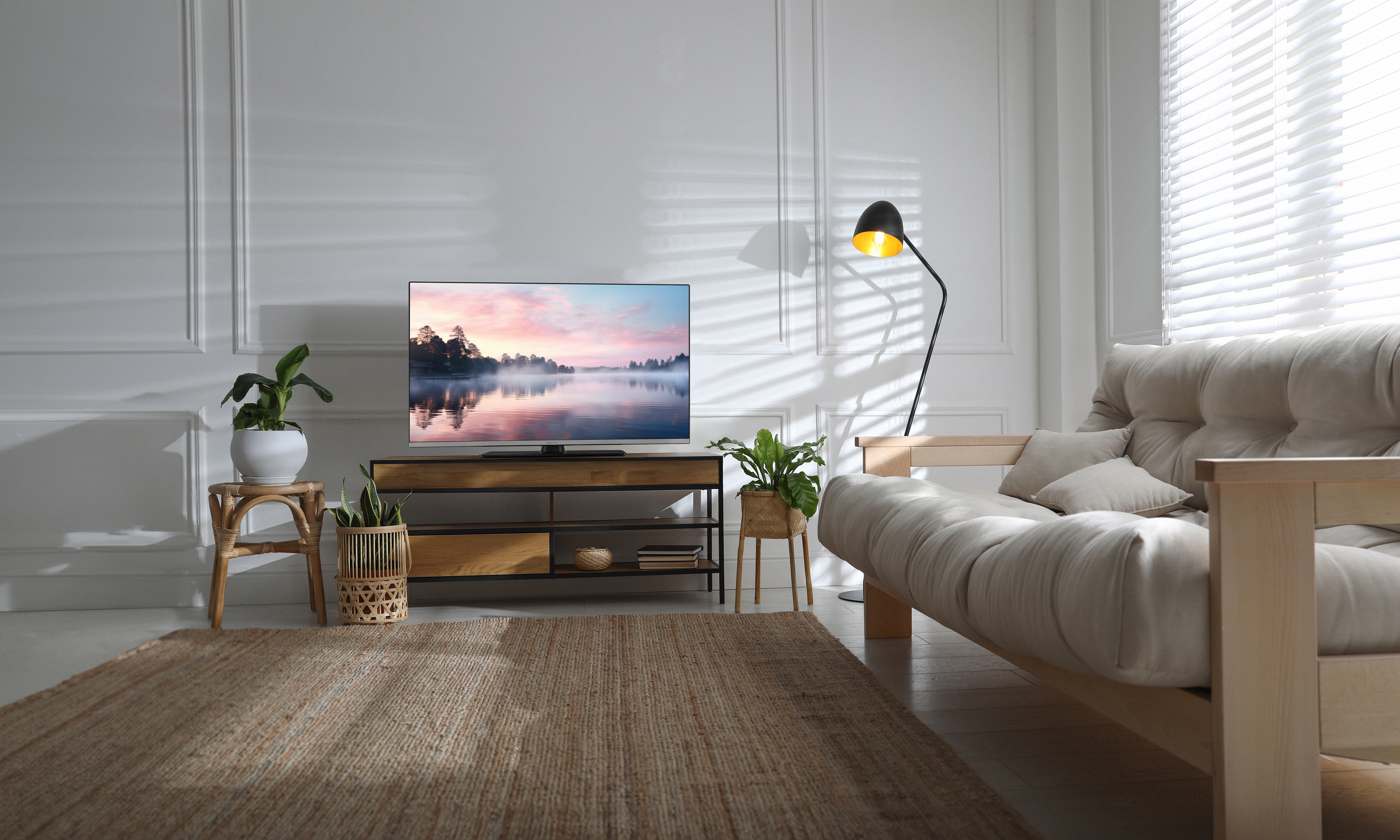 Ein Wohnzimmer mit Sofa, TV und Teppich. Sonnenlicht fällt durch Jalousien.