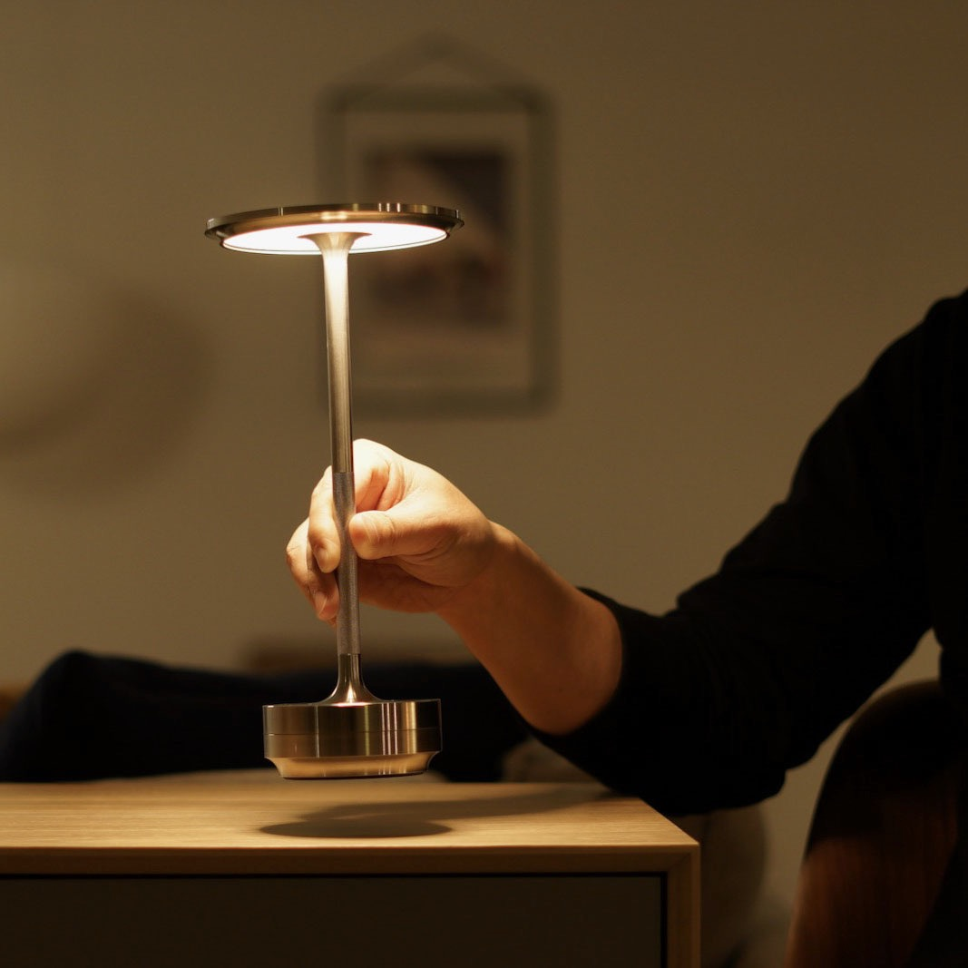 Person hält eine moderne, metallische Lampe mit rundem Lichtkopf über einem Holztisch.