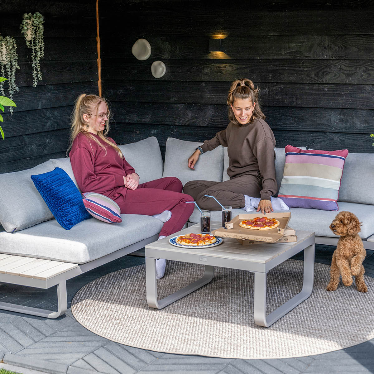 Two women sit on a couch eating pizza with a dog nearby.