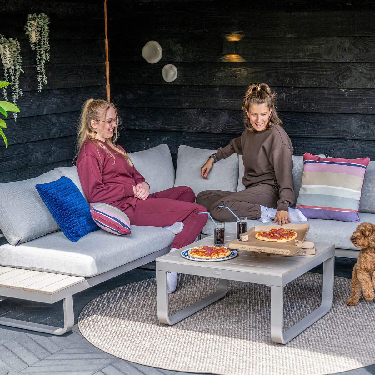 Two women sit on an outdoor couch with pizza and drinks. A dog sits beside them.