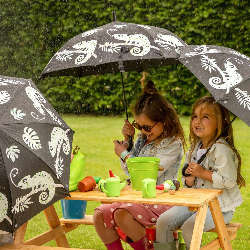 Zwei Mädchen sitzen im Regen unter Schirmen mit Chamäleons an einem Picknicktisch.