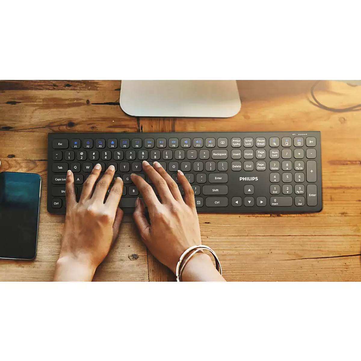 Manos escribiendo en un teclado Philips negro, junto a un smartphone.
