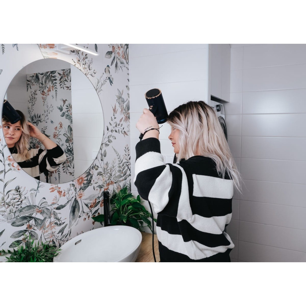 Kobieta z blond włosami suszy włosy suszarką w łazience z tapetą w kwiaty.