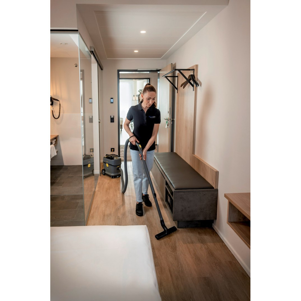 Kobieta odkurza drewnianą podłogę w pokoju hotelowym. Obok stoi odkurzacz.