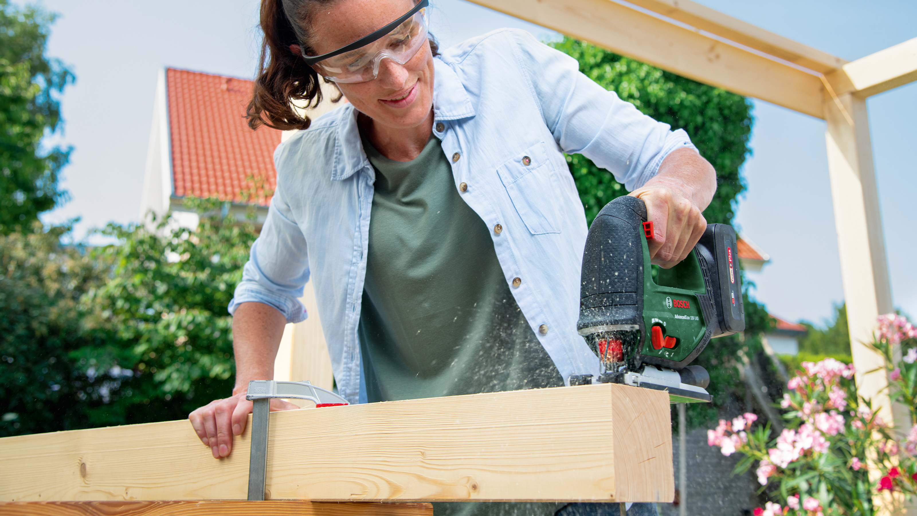 Frau mit Schutzbrille sägt Holz mit Stichsäge. Draußen, sonniger Tag.