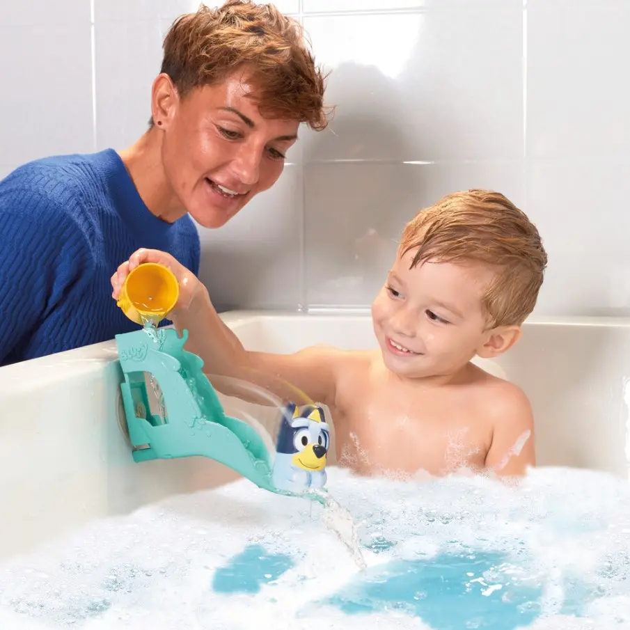 Una mujer vierte agua en un tobogán de juguete de baño azul para un niño sonriente en una bañera burbujeante.