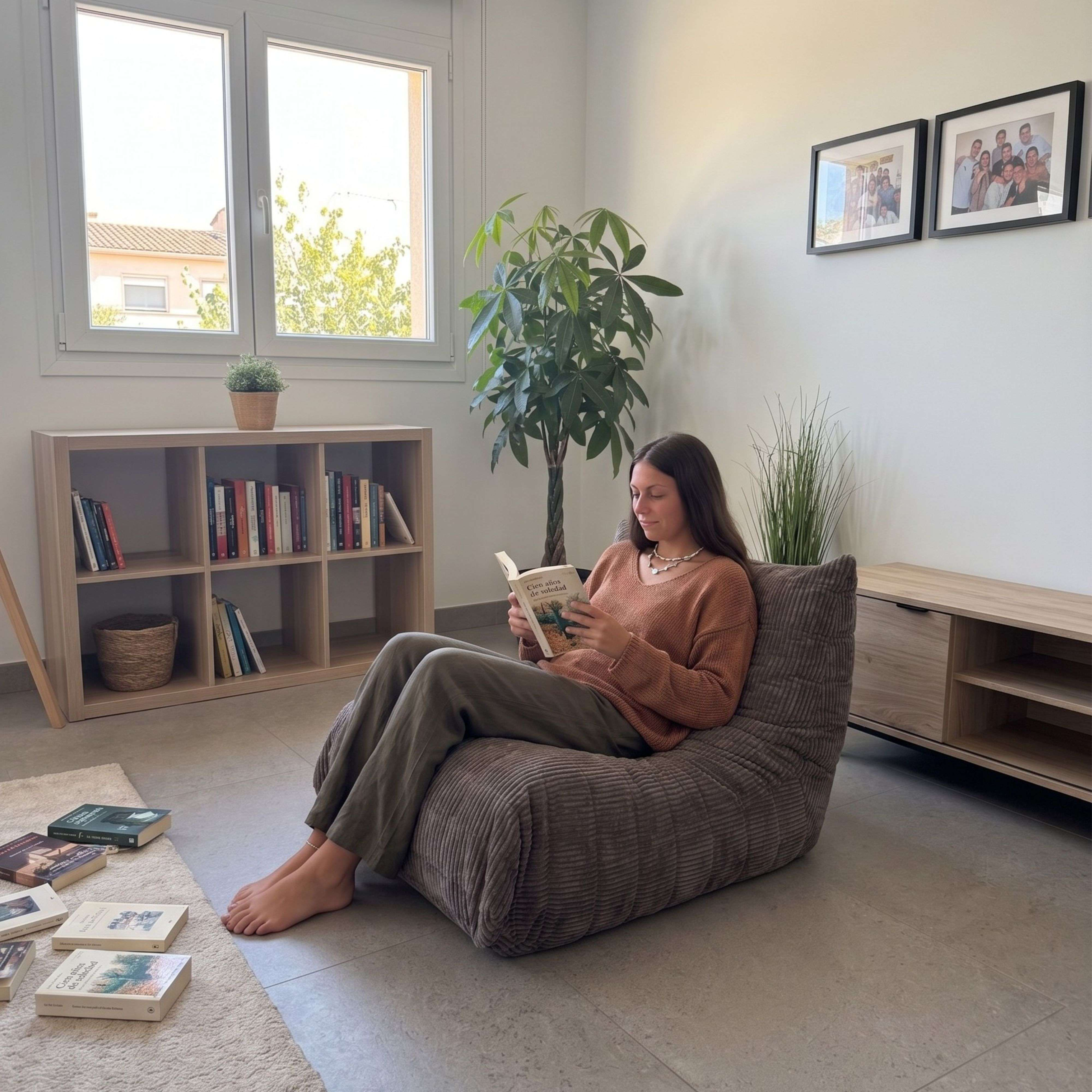 Mujer lee libro en puf marrón en sala, estantería al fondo.