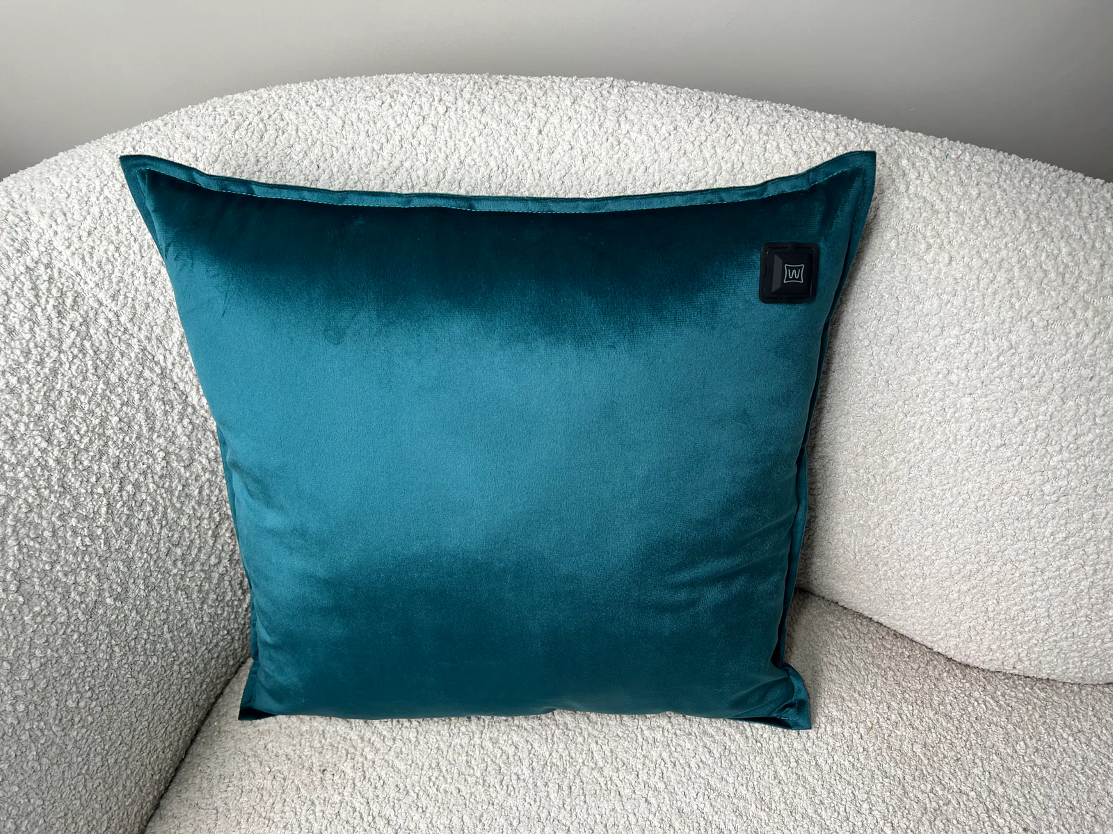 Ein türkiser Samtkissen liegt auf einem weißen Bouclé-Sofa. Ein schwarzes Etikett ist sichtbar.