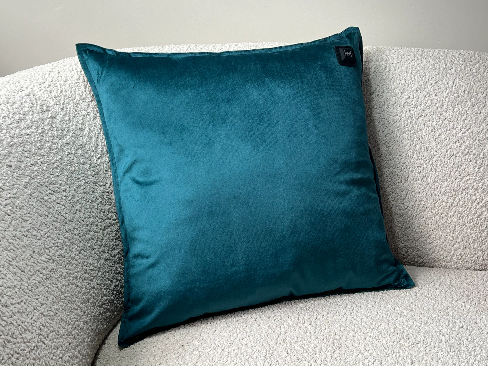 Ein petrolfarbenes Samtkissen mit Heizfunktion liegt auf einem hellgrauen Bouclé-Sofa.