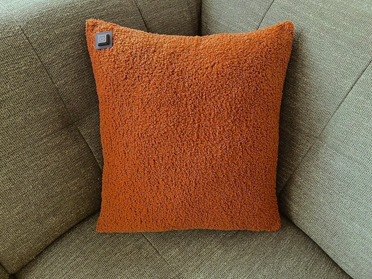 Ein orangefarbenes Kissen mit Teddyfell-Textur liegt auf einem grünen Sofa. Oben links ist ein Ein-/Ausschaltknopf.