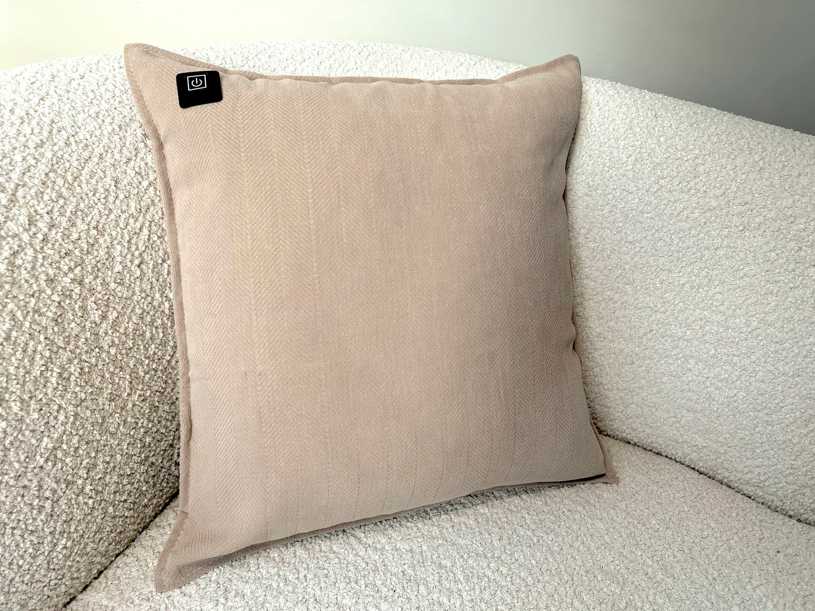 Ein beiger Kissen mit Fischgrätmuster auf einem cremefarbenen Bouclé-Sofa. Ein schwarzes Logo mit einem Power-Symbol.