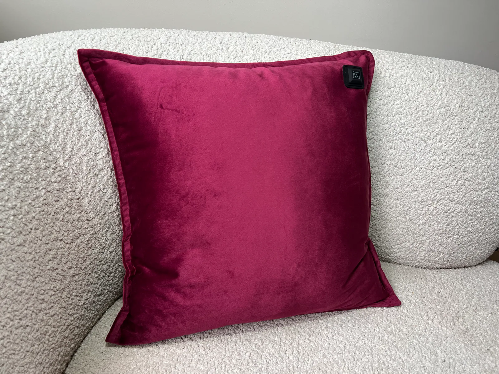 Ein dunkelrotes Samtkissen mit einem schwarzen Etikett liegt auf einem weißen Bouclé-Sofa.