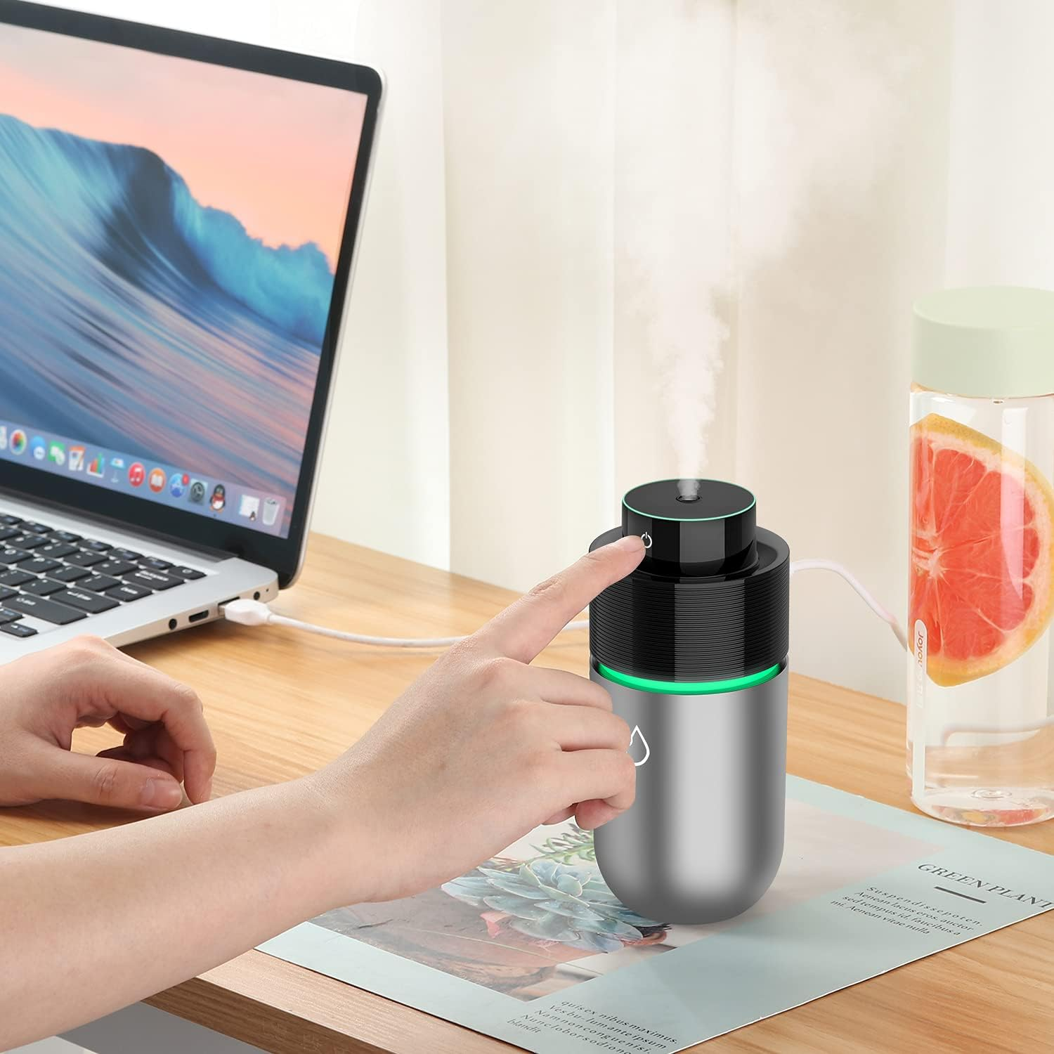 Ein Finger drückt auf einen silbernen Luftbefeuchter neben einem Laptop. Ein Wassernebel steigt auf.