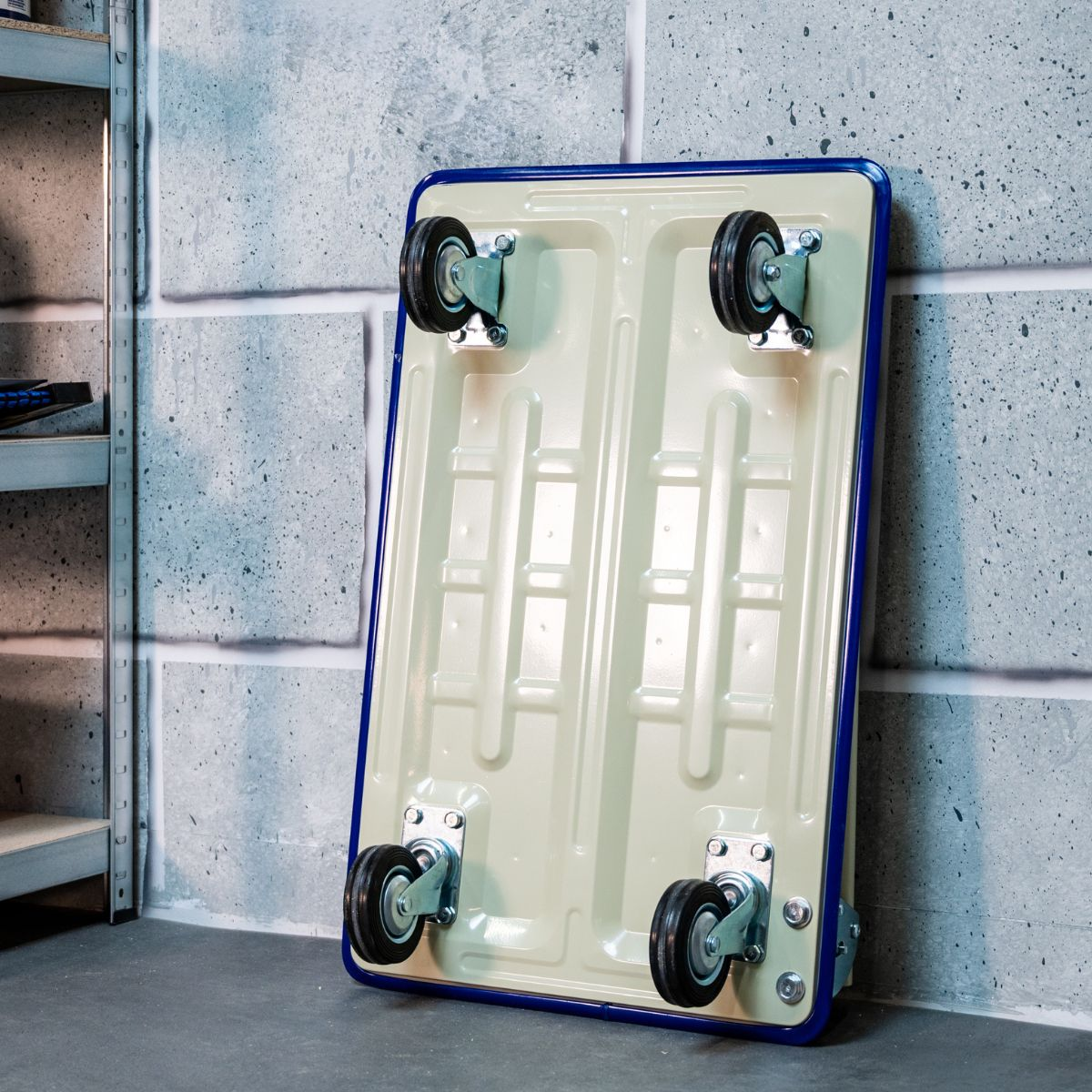 A cream dolly with blue trim and four wheels stands against a concrete wall.
