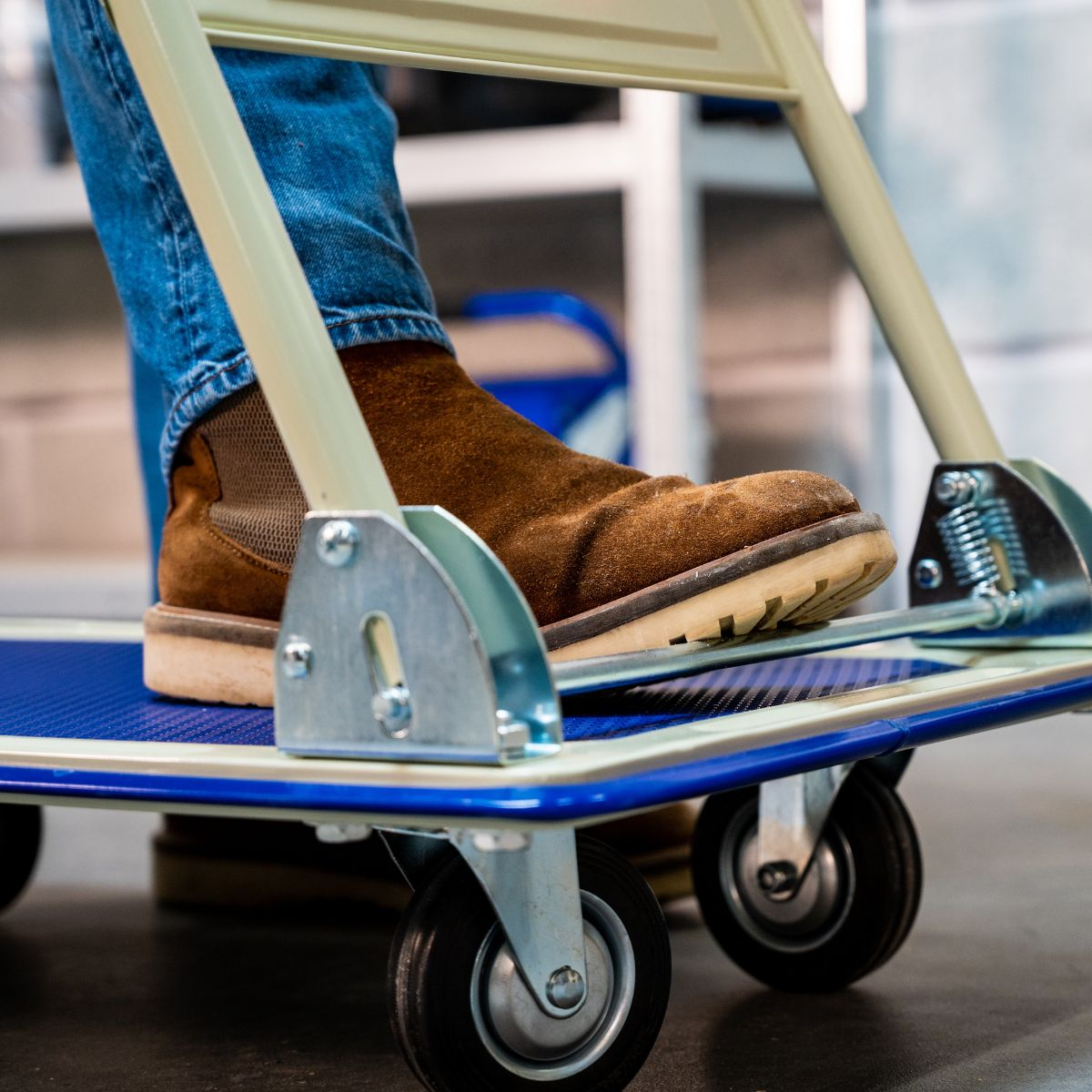 A person in blue jeans and a brown boot stands on a blue dolly with silver wheels.