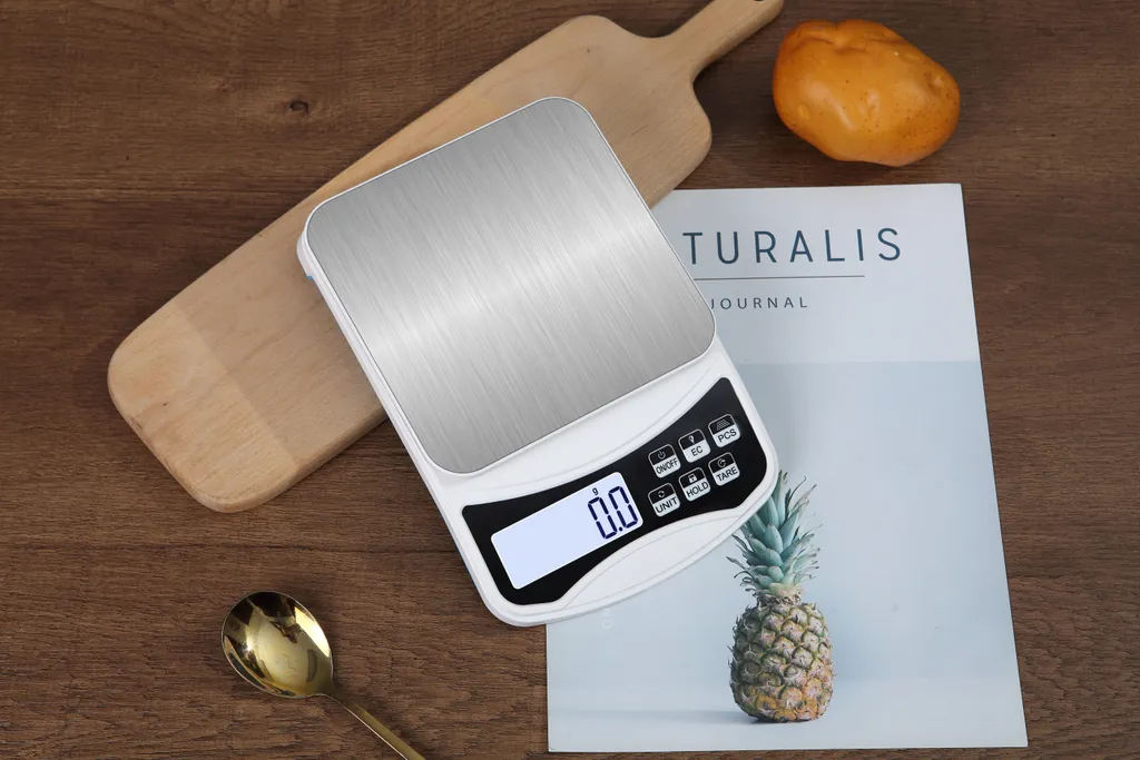Eine digitale Küchenwaage mit Edelstahlplatte, daneben ein Holzbrett, Löffel, Ananas und ein Buch.