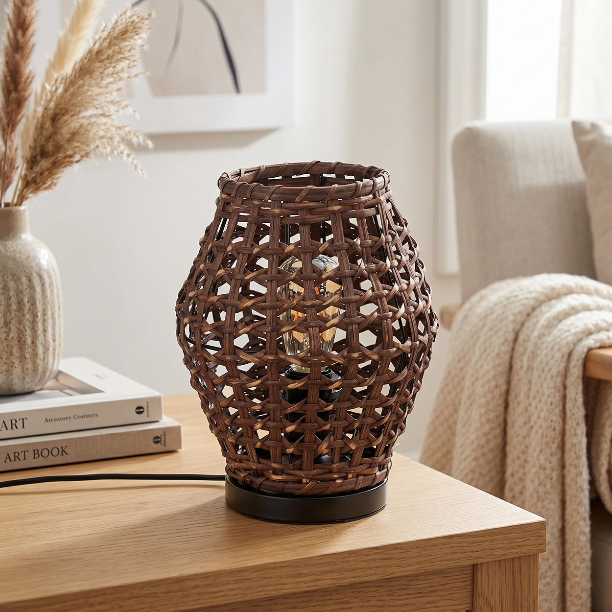 Braune Weidenlampe mit Glühbirne auf einem Holztisch, neben einem Vase mit Gräsern.