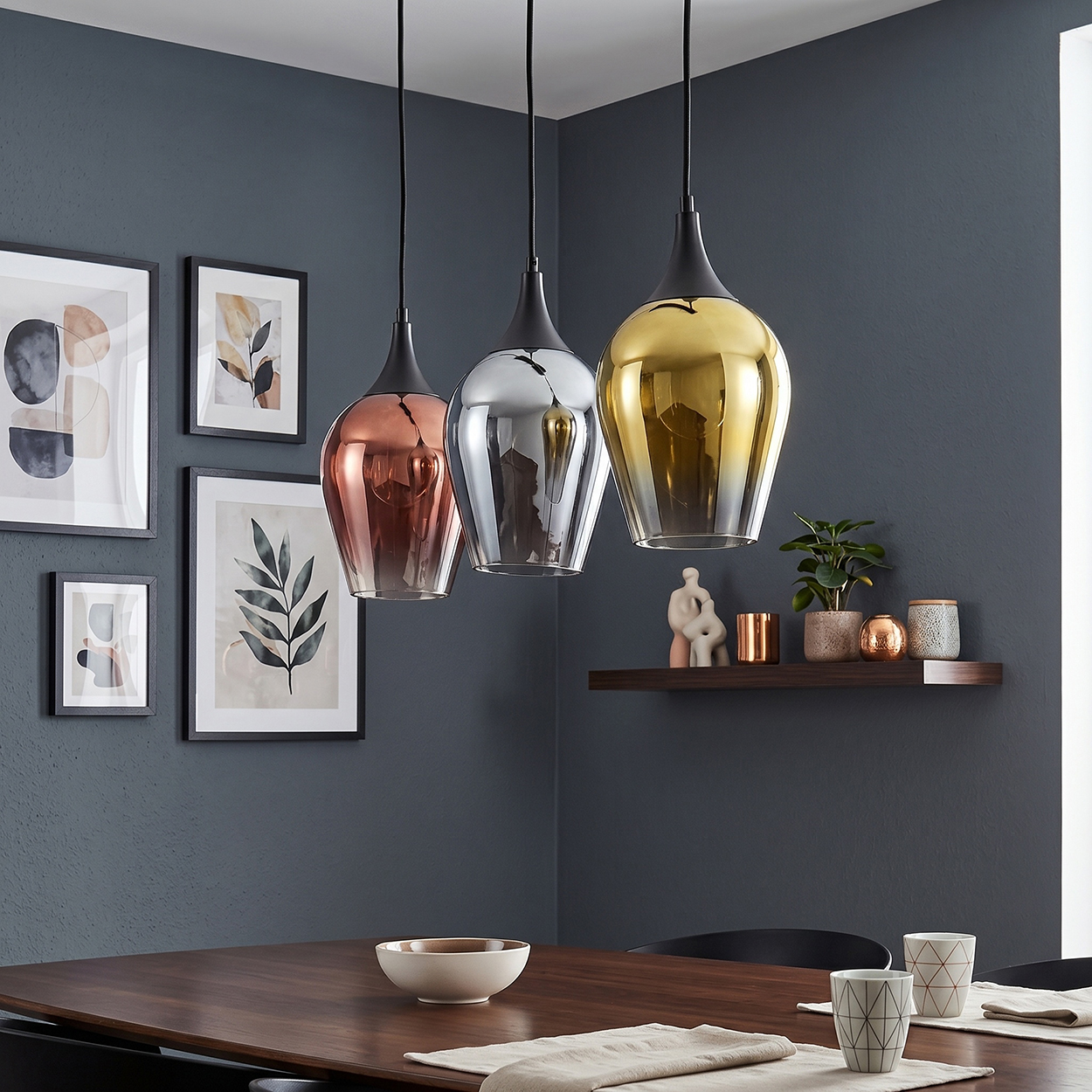 Drei Hängeleuchten in Kupfer, Silber und Gold über einem Holztisch.