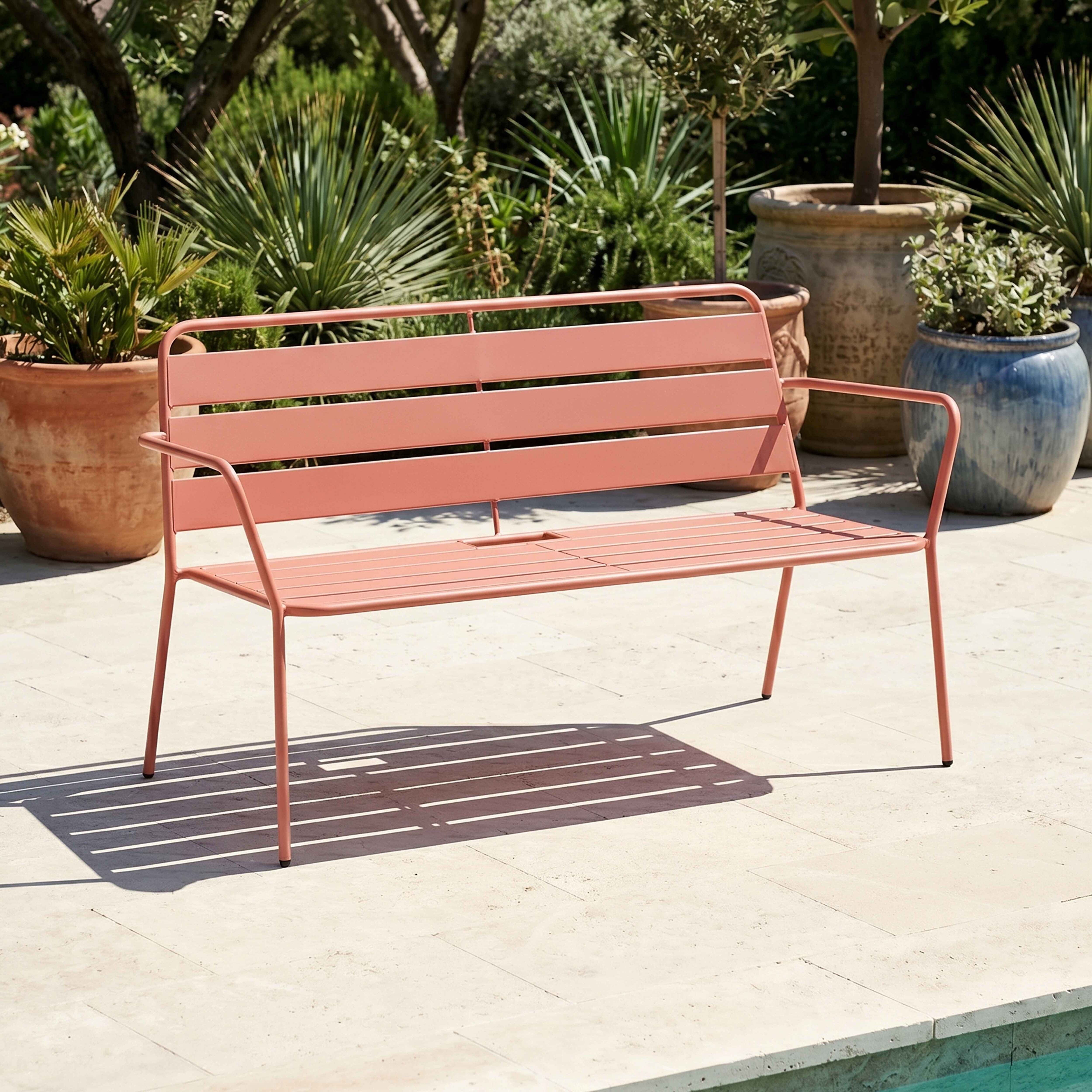 Ein korallenrotes Gartensofa steht auf einer hellen Steinterrasse neben großen Blumentöpfen.
