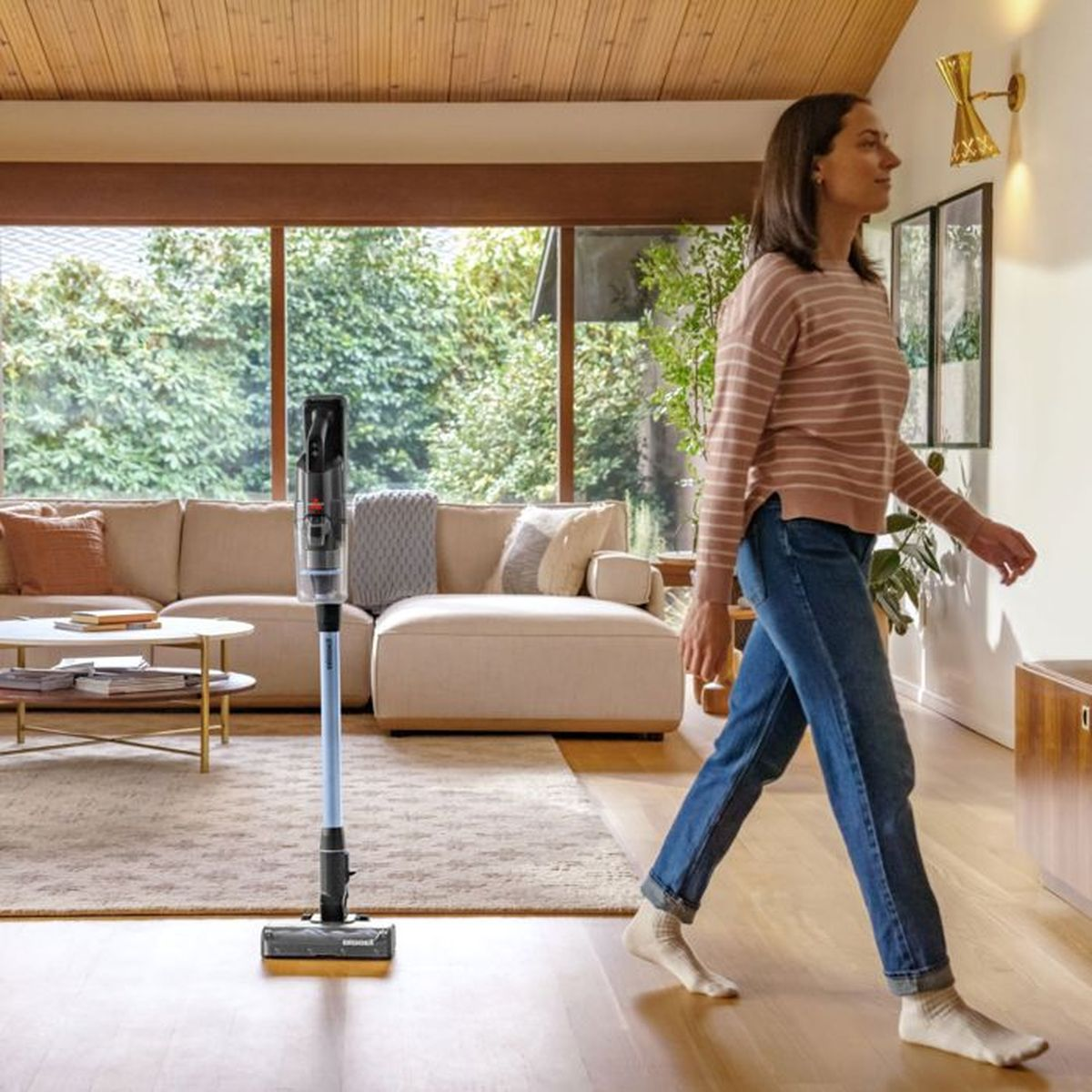 Eine Frau in gestreiftem Pullover und Jeans geht im Wohnzimmer an einem Staubsauger vorbei.