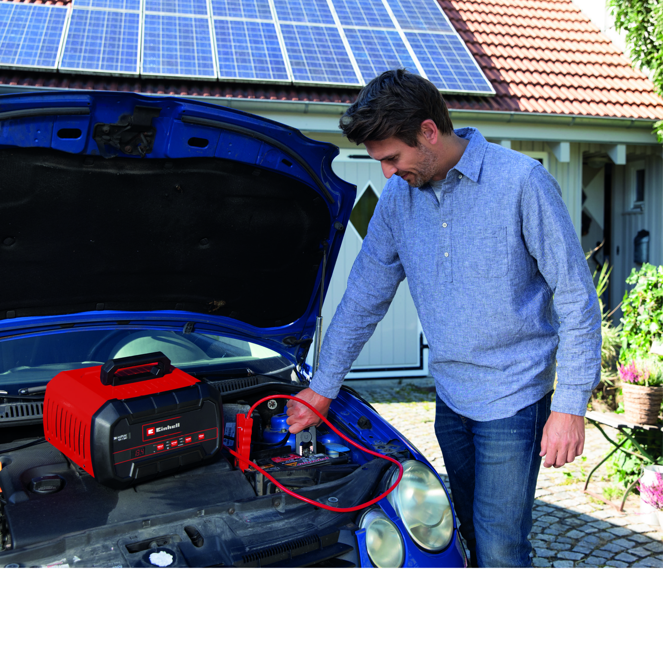 Mann mit Starthilfegerät am offenen blauen Auto, Solarzellen auf dem Dach.